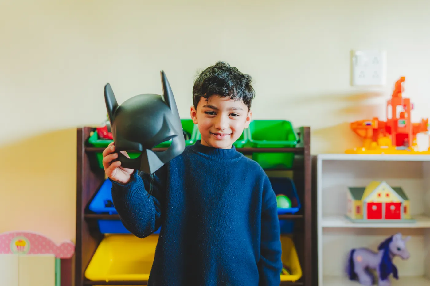 A boy holding up a superhero mask in the toy room